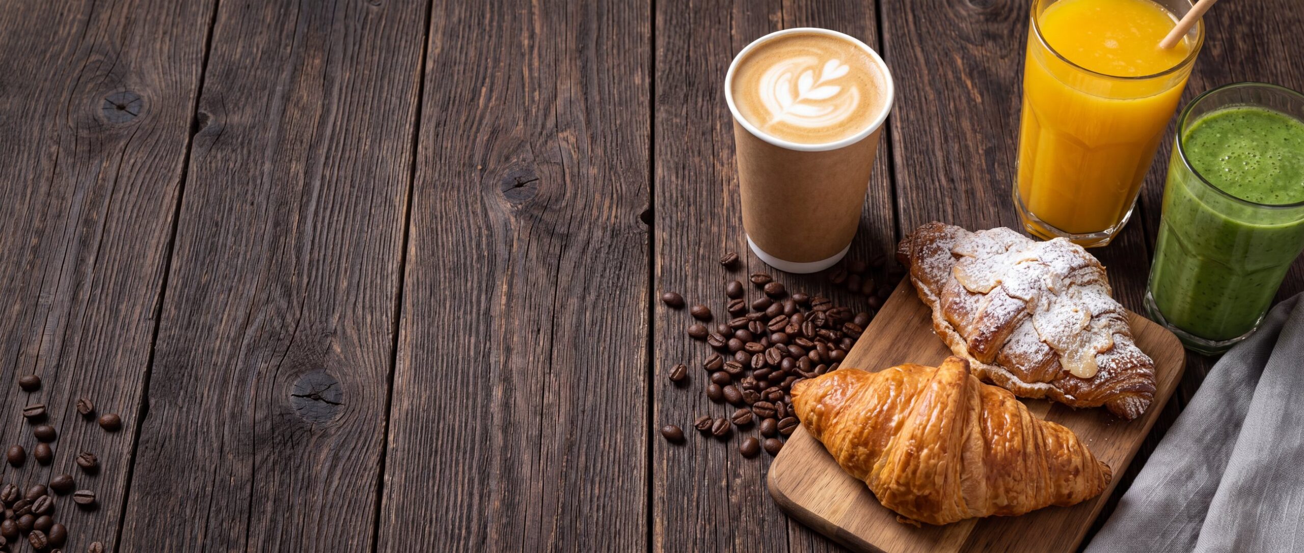 A flat white in a kraft cup, croissants, orange juice and a green smoothie on dark wood
