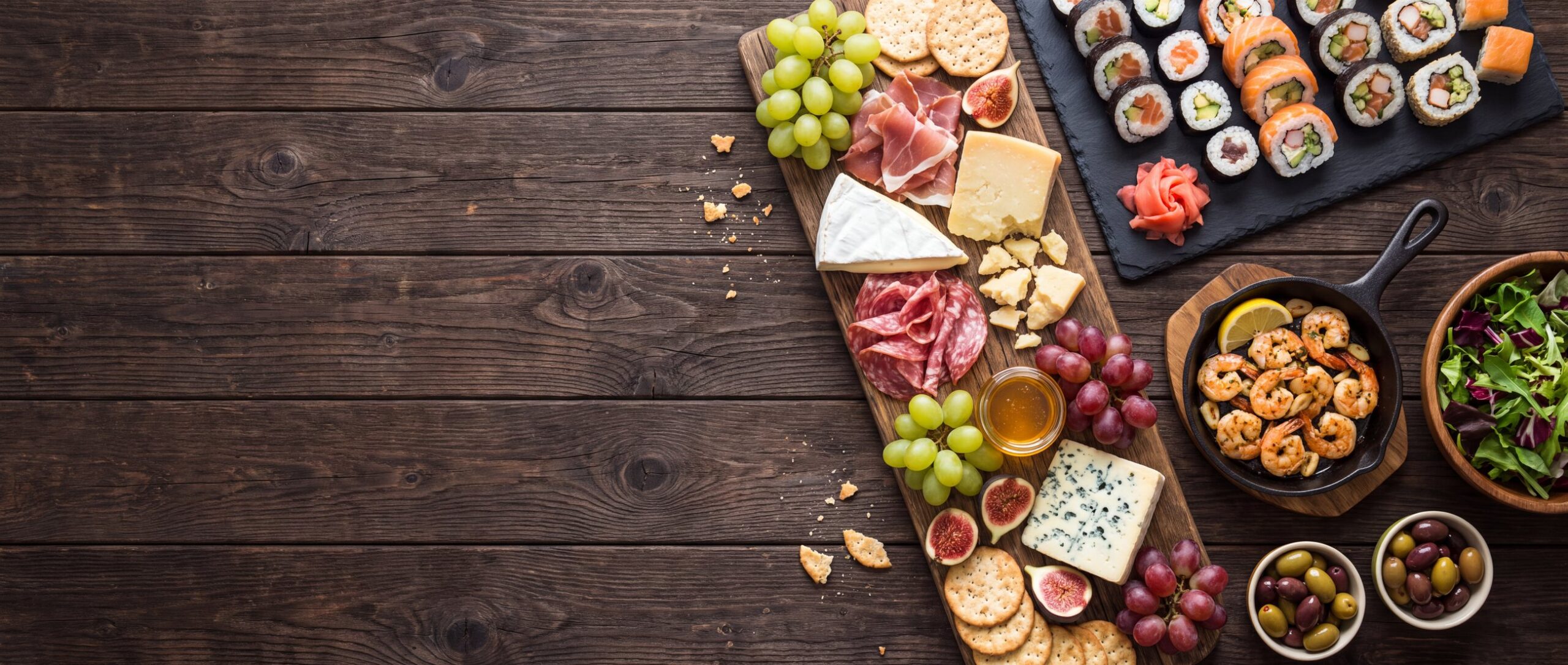 A catering spread: cheese board, sushi, garlic shrimp and salad on dark wood