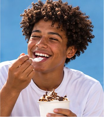 A laughing young man with a cup of frozen yogurt, against a blue wall