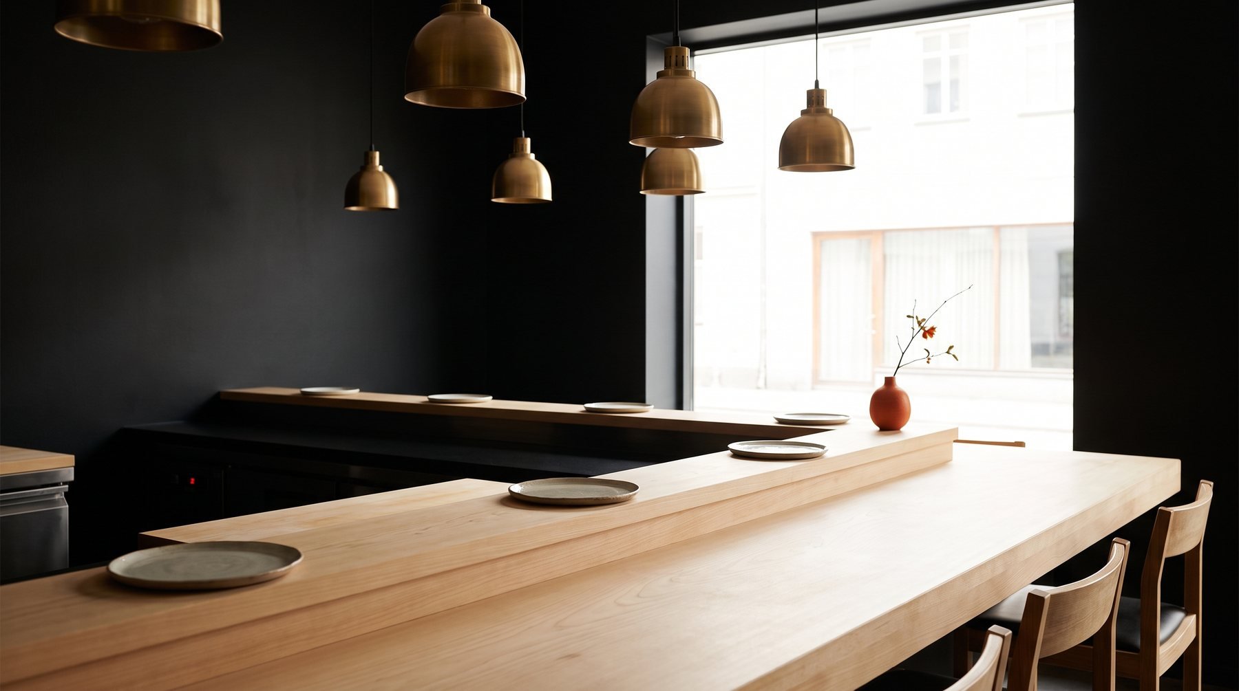 A long pale wooden counter with grey plates and wooden chairs under brass pendant lamps