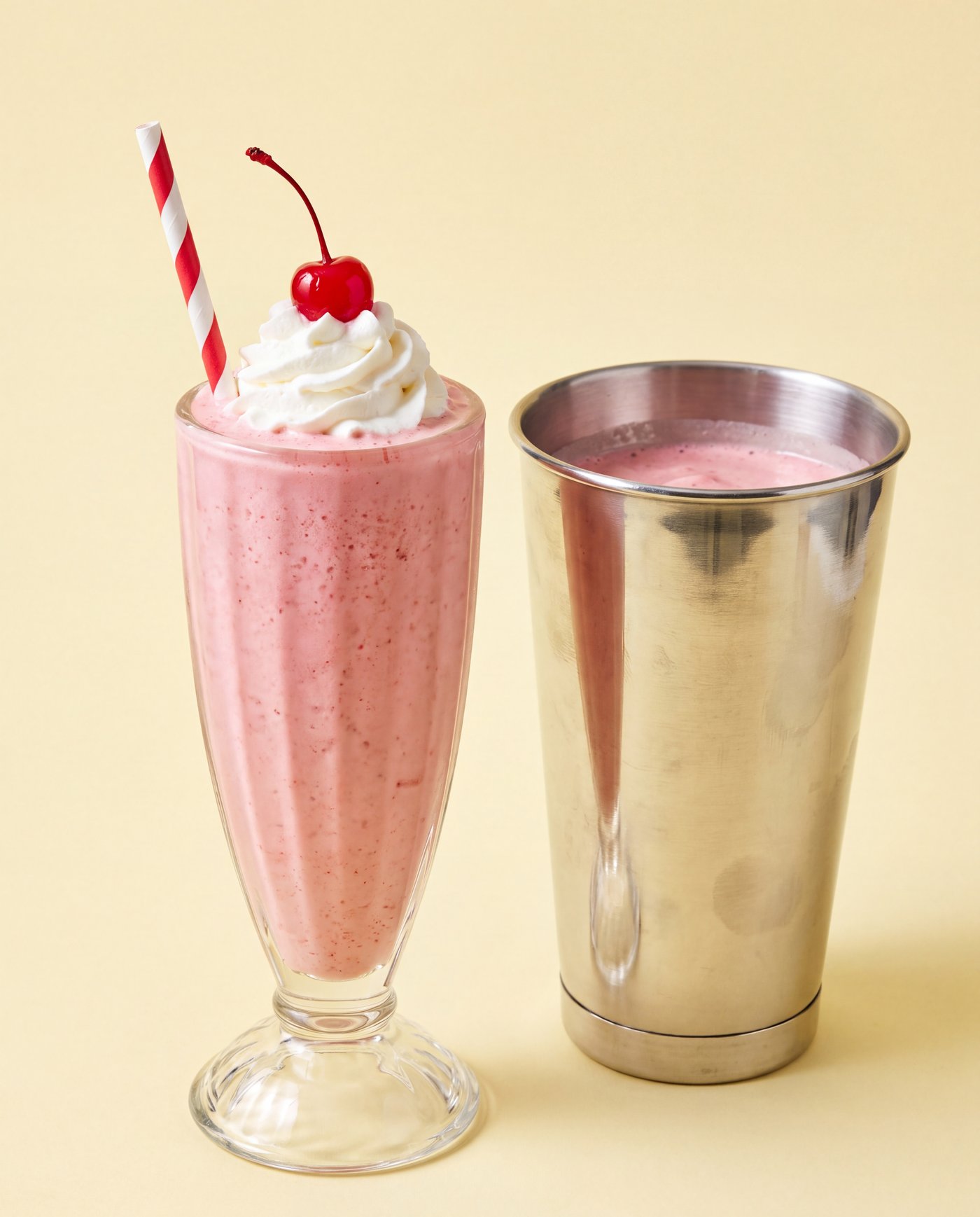 A tall strawberry frozen custard shake with whipped cream and the steel tin beside it