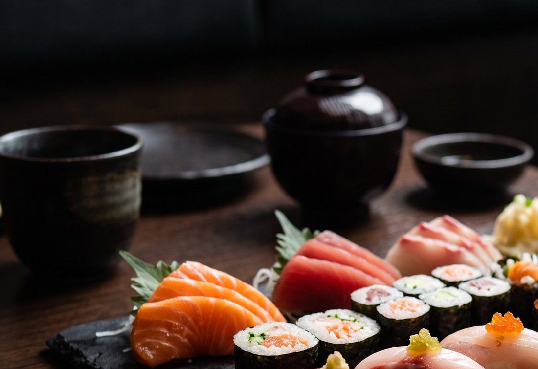 A lidded black soup bowl, a stoneware cup and small dishes on a dark table beside a slate of sushi