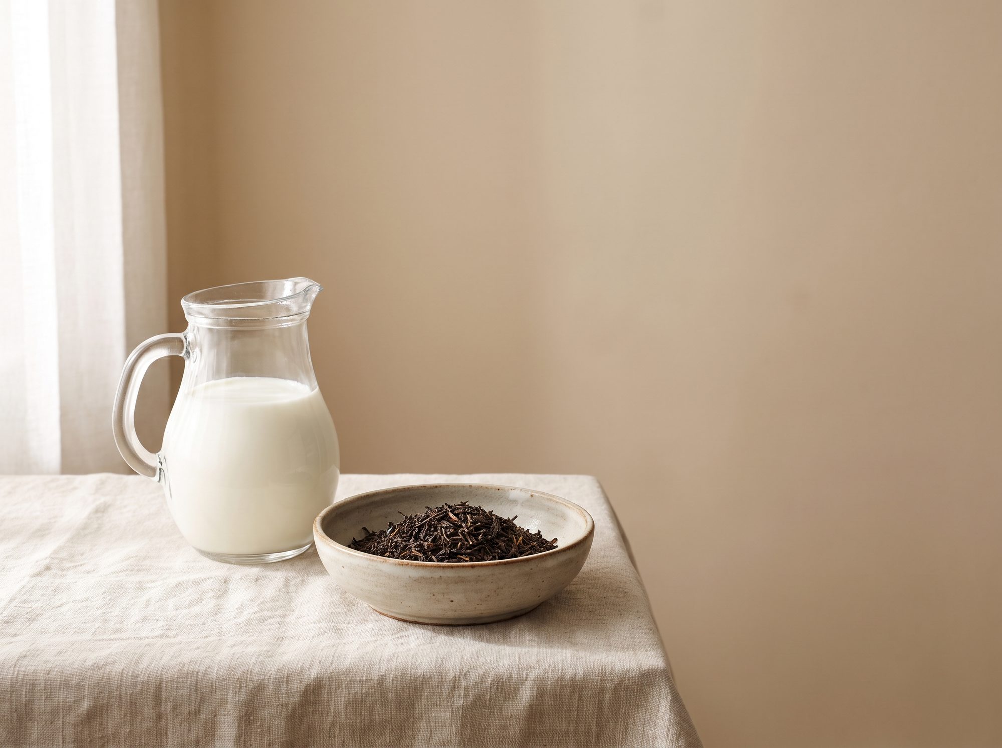 A glass jug of whole milk beside a dish of loose-leaf tea in window light