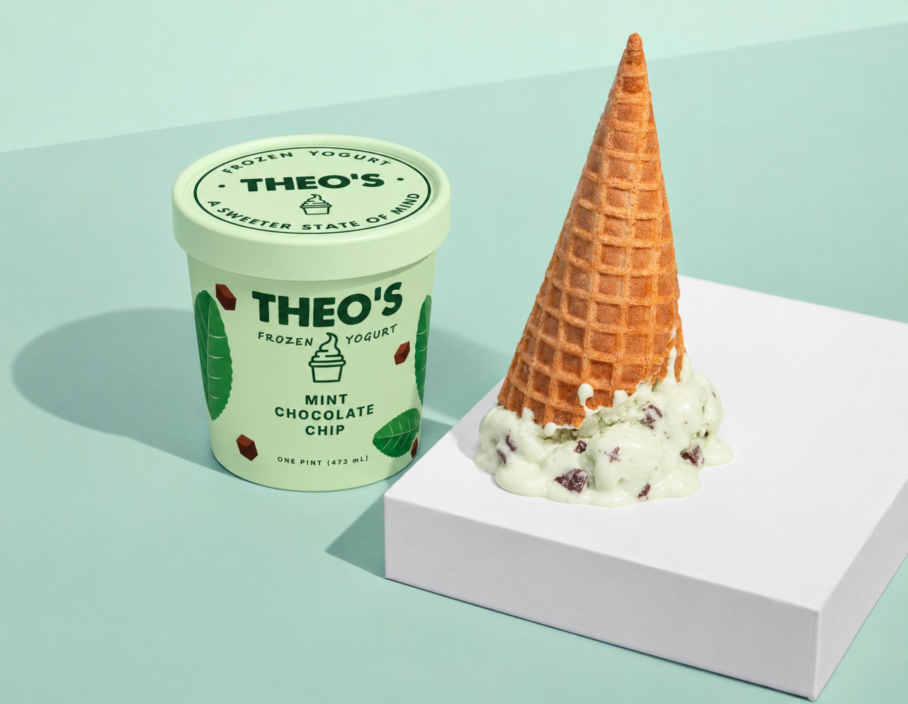 A Theo’s Mint Chocolate Chip pint beside an upturned waffle cone melting into mint chip on a white block