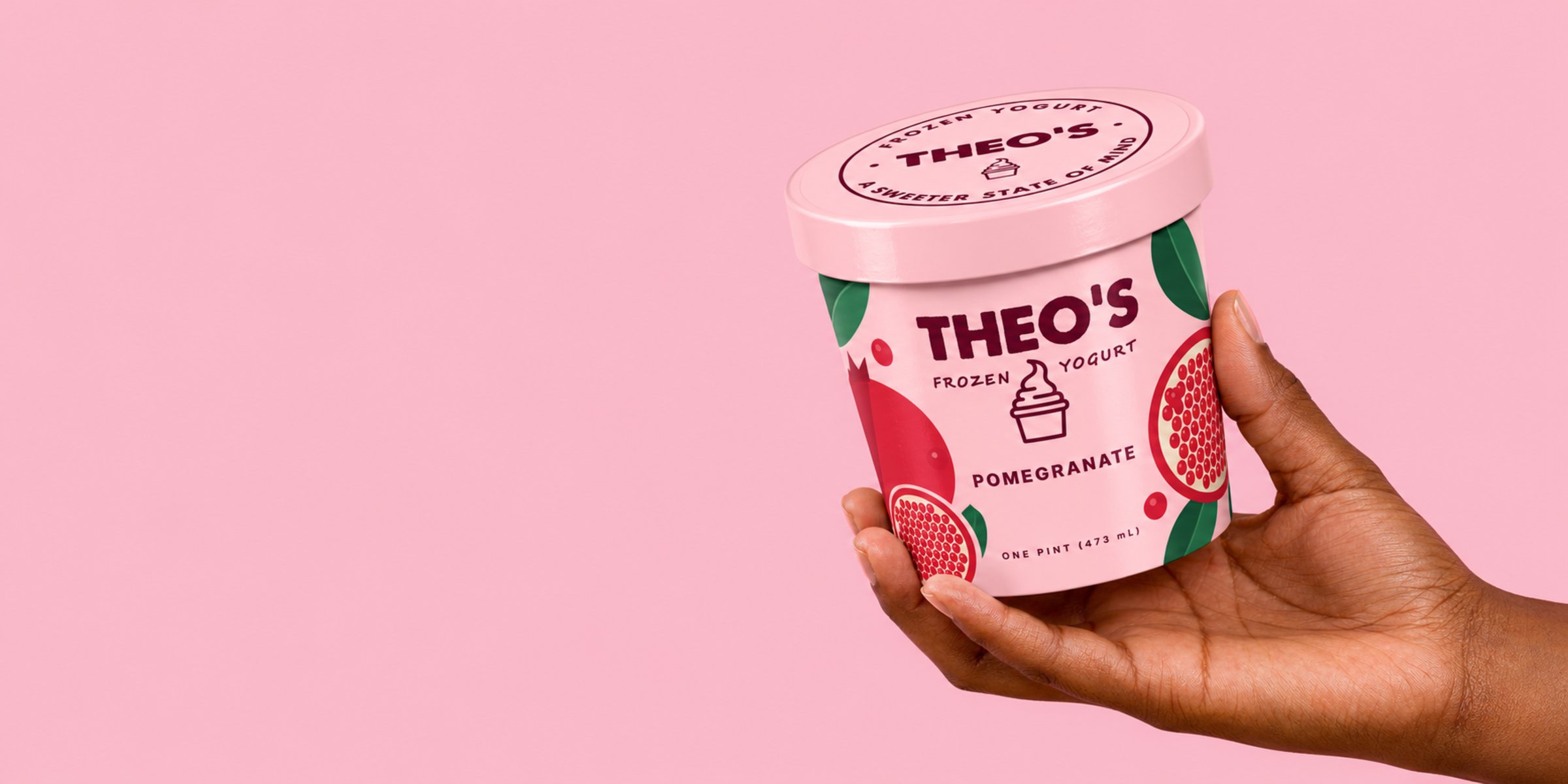 A hand holds up a Theo’s Pomegranate pint against a pink wall
