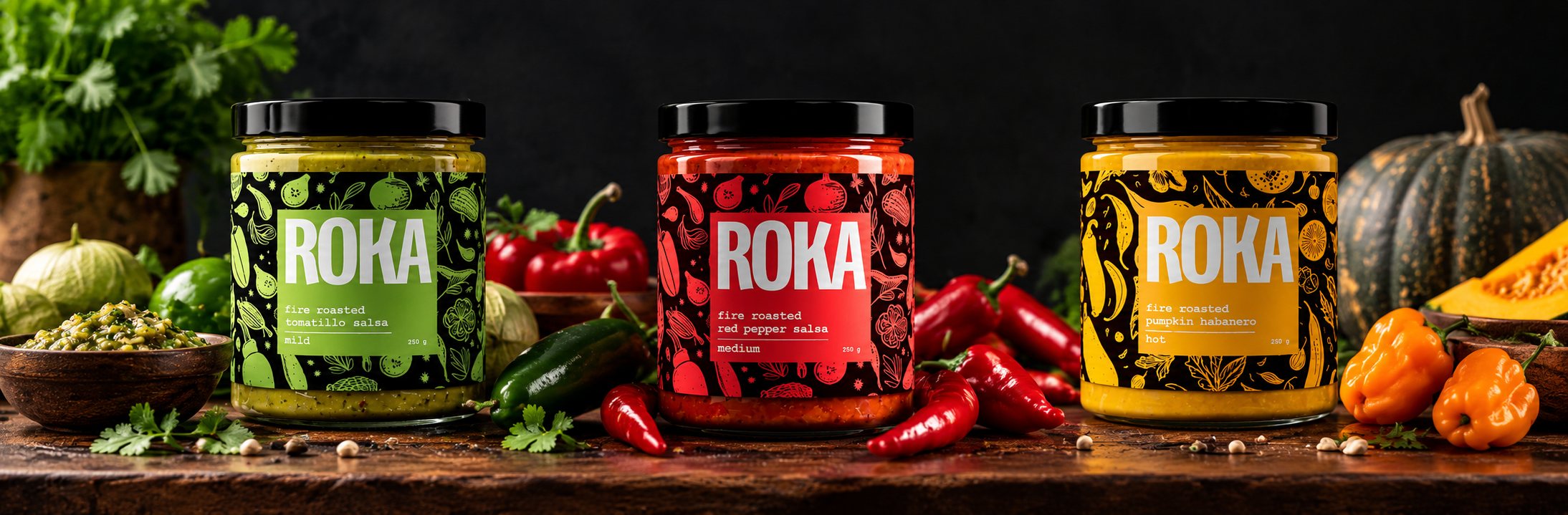 Three ROKA jars on a wooden table among tomatillos, red peppers, habaneros and a pumpkin: tomatillo salsa (mild), red pepper salsa (medium) and pumpkin habanero (hot), 250 g each