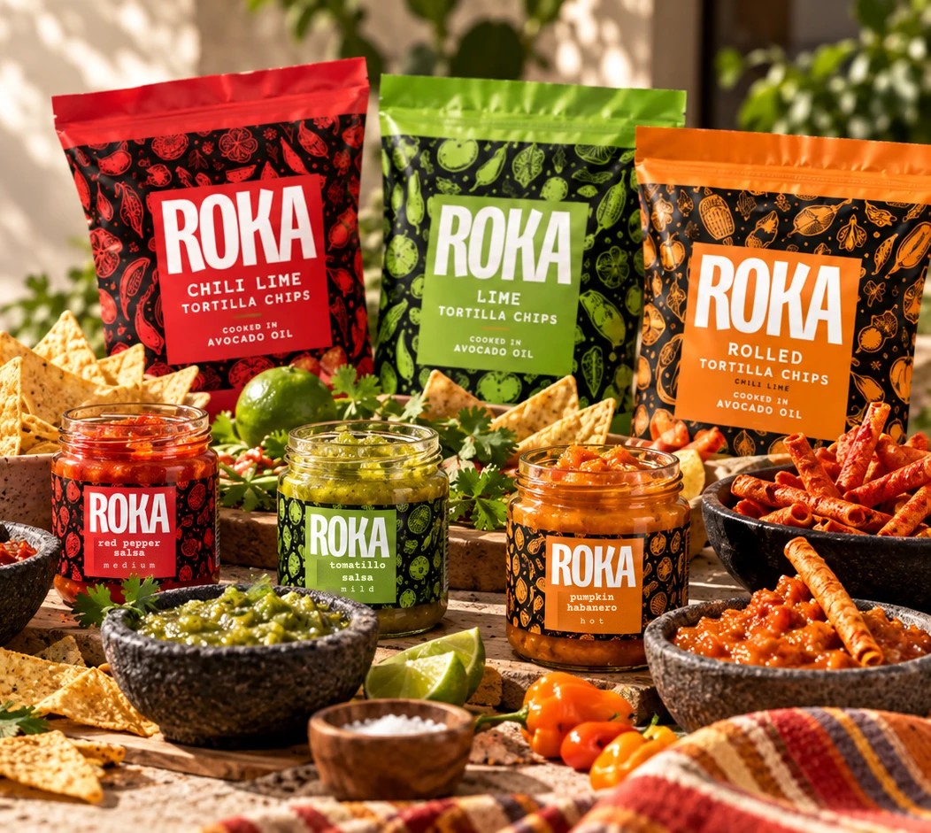 Three bags of ROKA tortilla chips, chili lime in red, lime in green and rolled chili lime in orange, standing behind the three salsa jars, with bowls of salsa, chips, limes and peppers on a sunny table