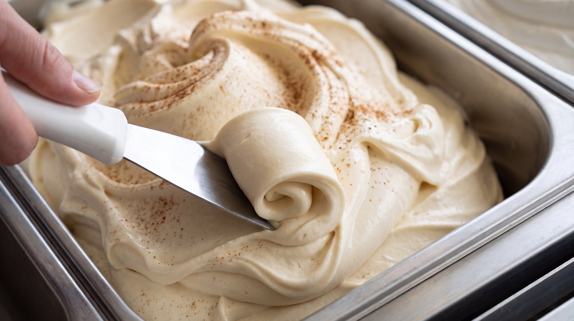 A hand working a steel spade through a deep pan of fresh frozen custard dusted with cinnamon