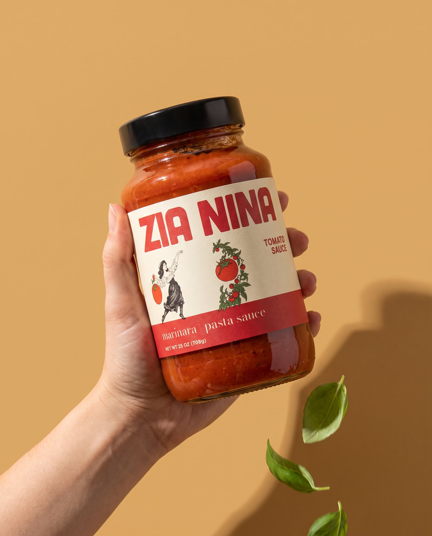 A hand holding up a jar of Zia Nina Marinara against a golden wheat backdrop, basil leaves falling past